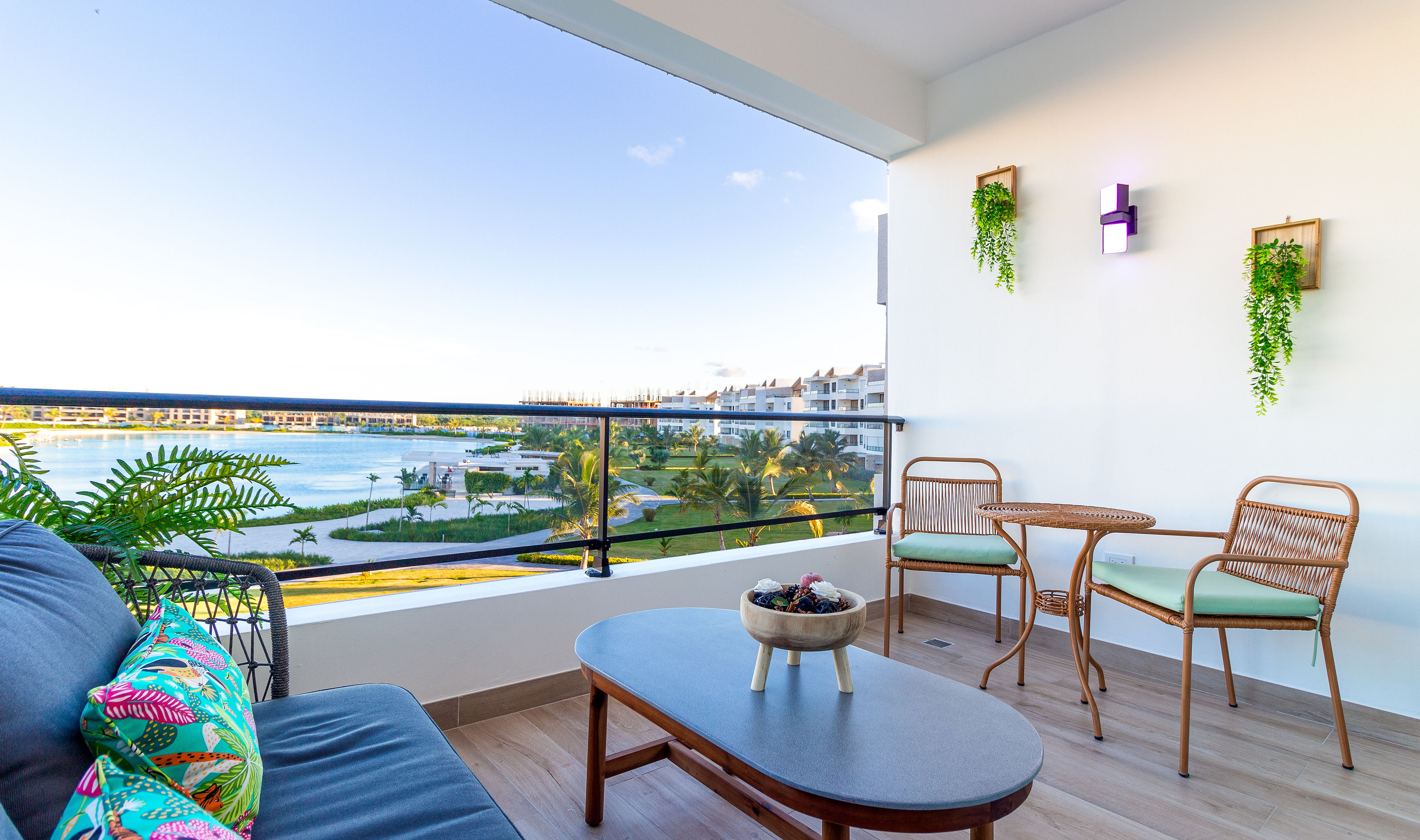 A terrace featuring a comfortable grey sofa adorned with a colorful tropical pillow and a round wooden coffee table. Two stylish woven chairs with green cushions surround the table. The terrace overlooks a serene body of water with palm trees and residential buildings in the background, framed by a bright blue sky.