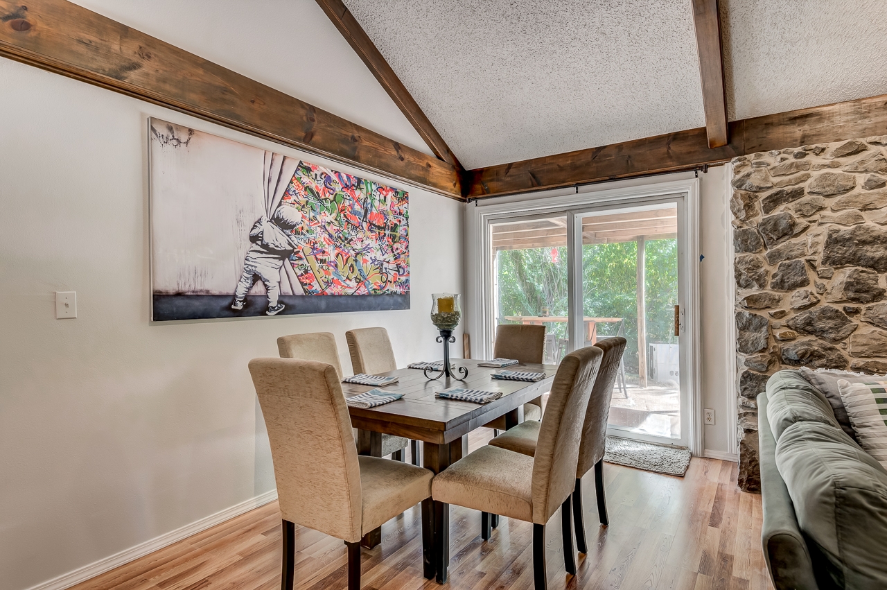 A cozy dining room featuring a wooden table surrounded by six upholstered chairs. The room has rustic wooden beams on the ceiling and a large colorful artwork on the wall. There are large glass doors leading to a deck, allowing natural light to fill the space. The floor is made of light-colored wood, complementing the warm tones of the room.