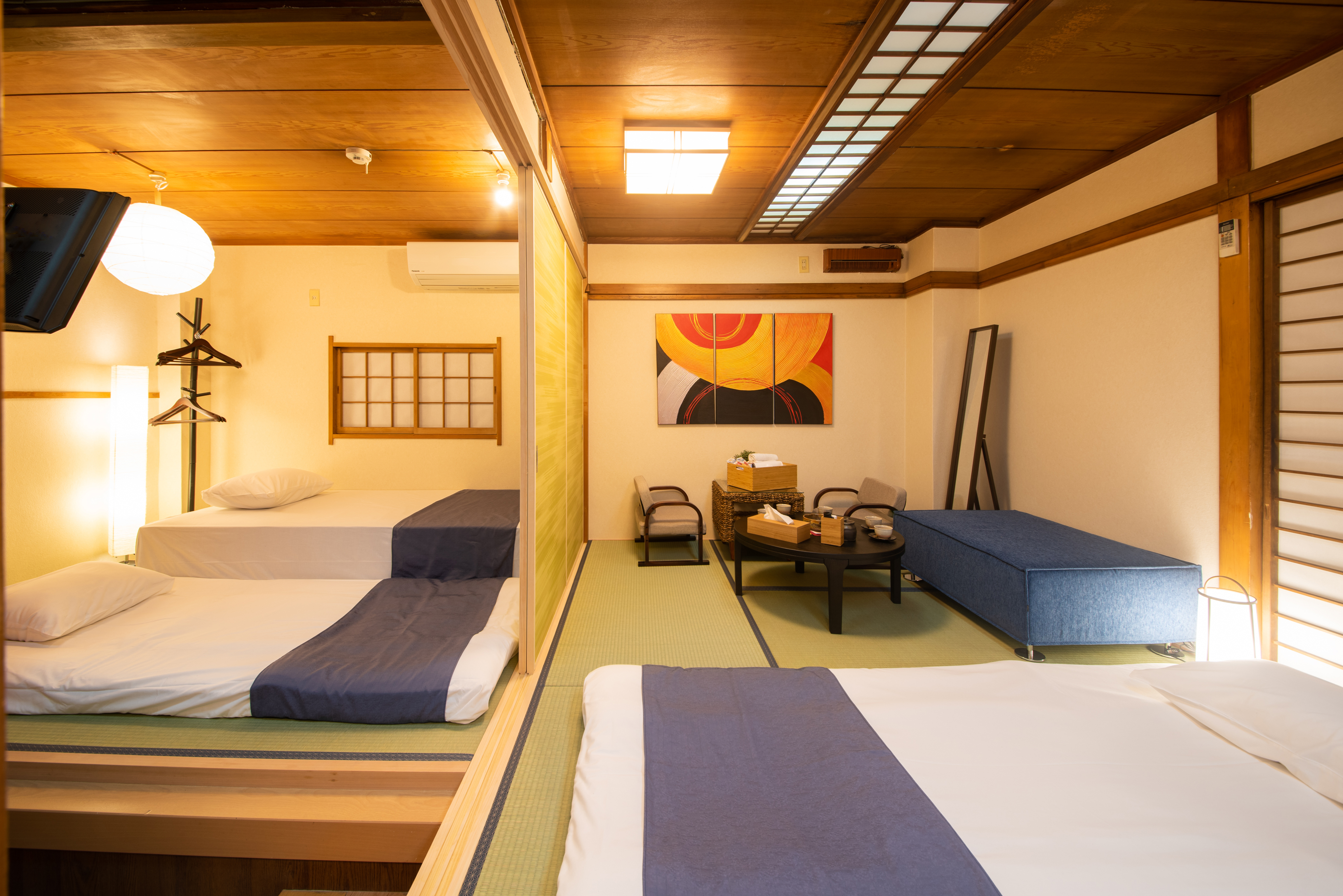A cozy bedroom featuring two beds with white linens and blue runners. The room has traditional Japanese decor with wooden beams, tatami mat flooring, and a decorative painting on the wall. A small table with chairs is situated in the center, alongside a blue sofa. Natural light floods through the window, enhancing the warm ambiance of the space.