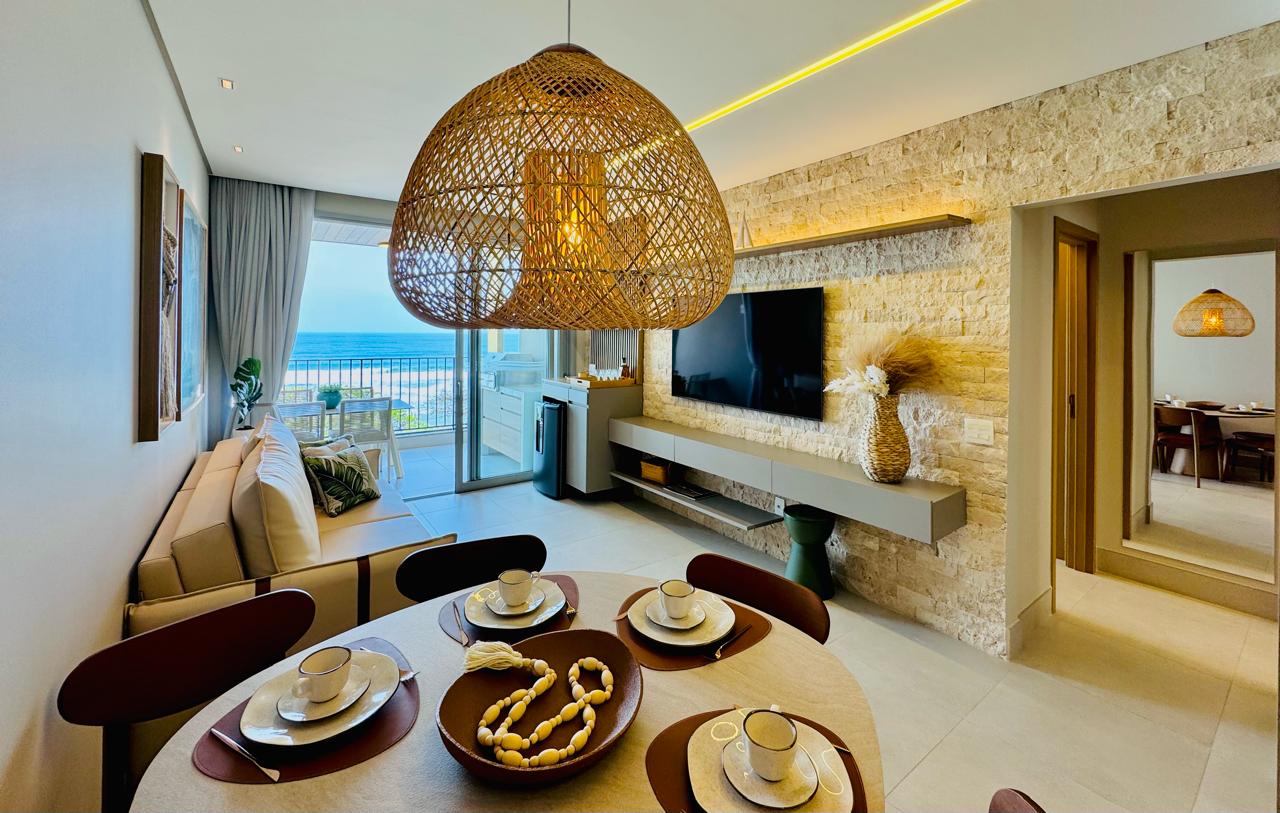 A bright and airy dining area in a beachfront apartment in Brazil, featuring a round wooden table set for four with elegant dishware, a stylish woven pendant light overhead, and large glass doors leading to a balcony with ocean views.