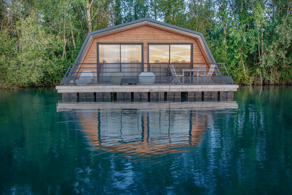 A modern wooden cabin with a sloped roof is situated on stilts over clear blue water, surrounded by lush greenery. The cabin features large glass windows and a balcony with outdoor seating, reflecting beautifully in the water below.