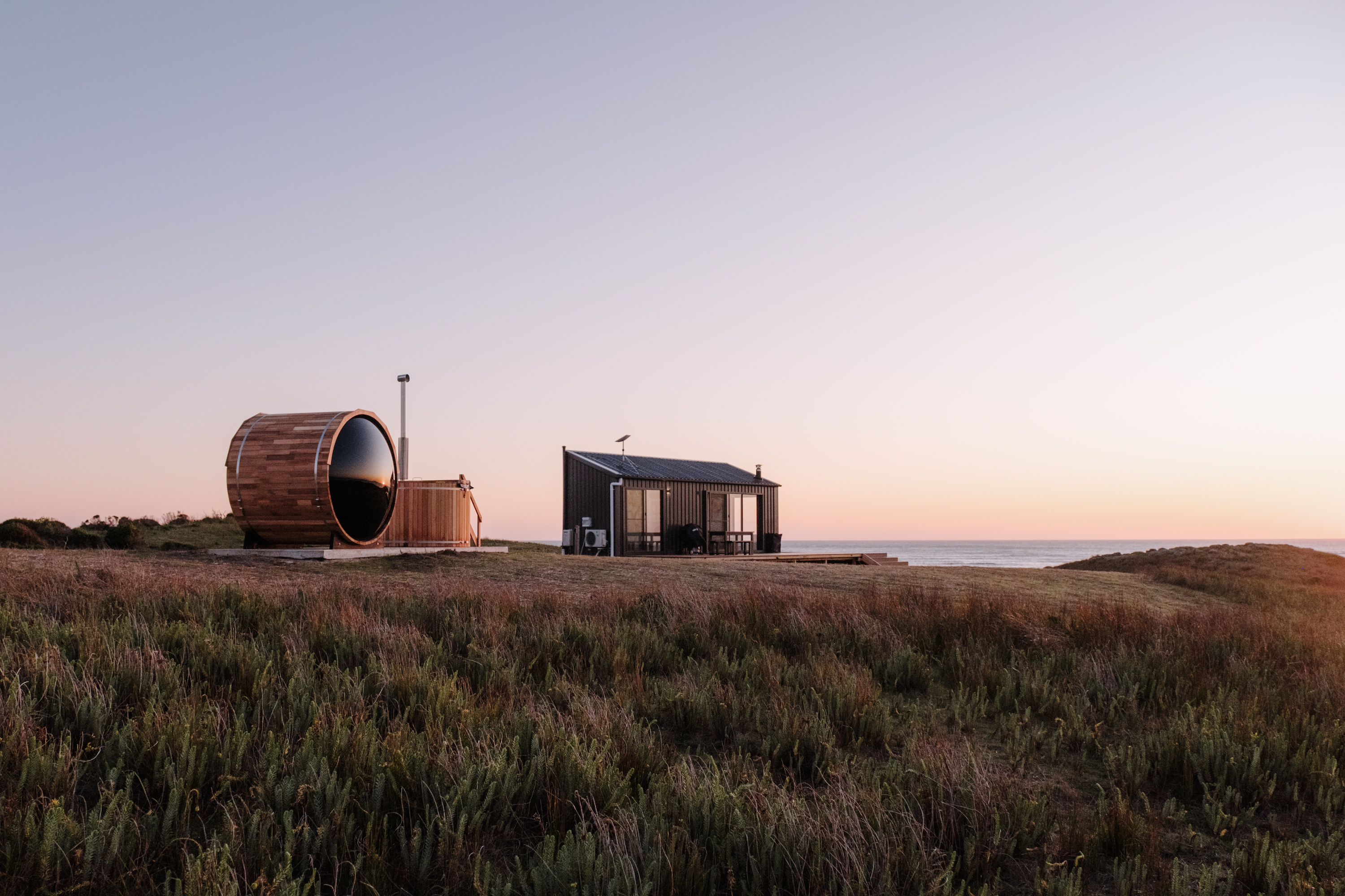 A tranquil seaside scene featuring a modern black cabin and a wooden hot tub with a rounded design, set against a serene sunset sky. The cabin is situated on a grassy area overlooking the ocean, with gentle hills in the background.