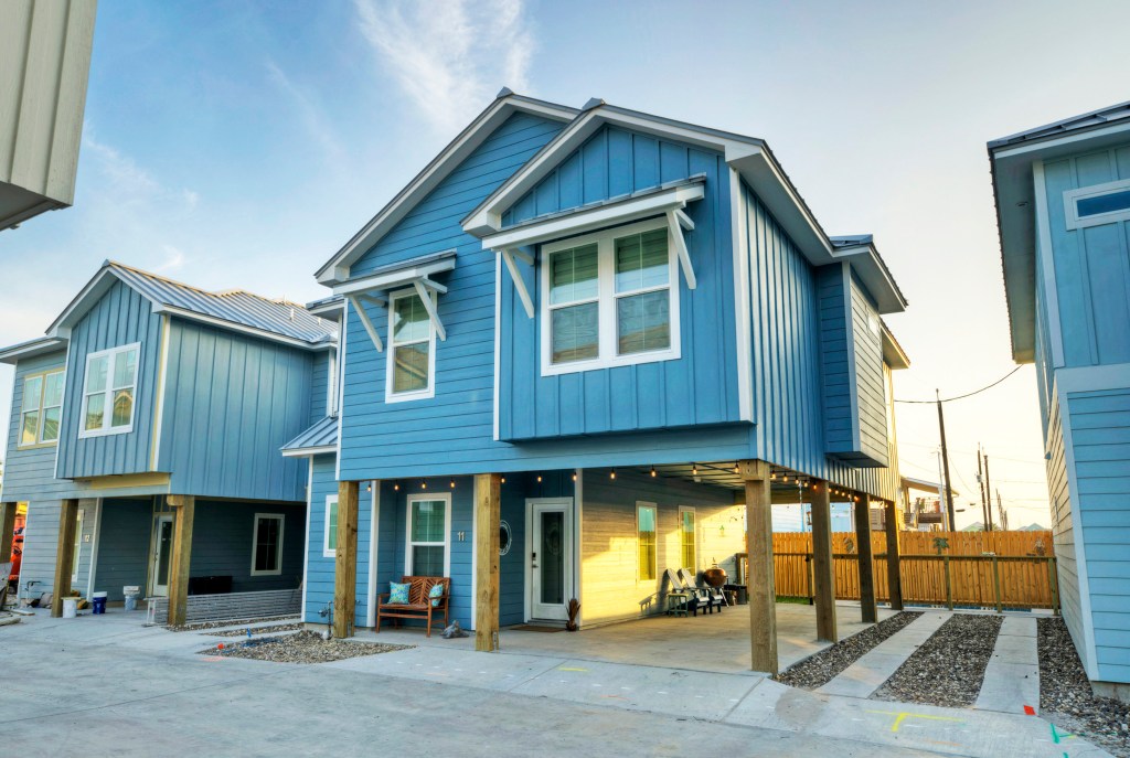 A modern blue two-story house with a metal roof, situated on stilts. The house features large windows and a covered porch area with seating. Gravel landscaping surrounds the base of the house.