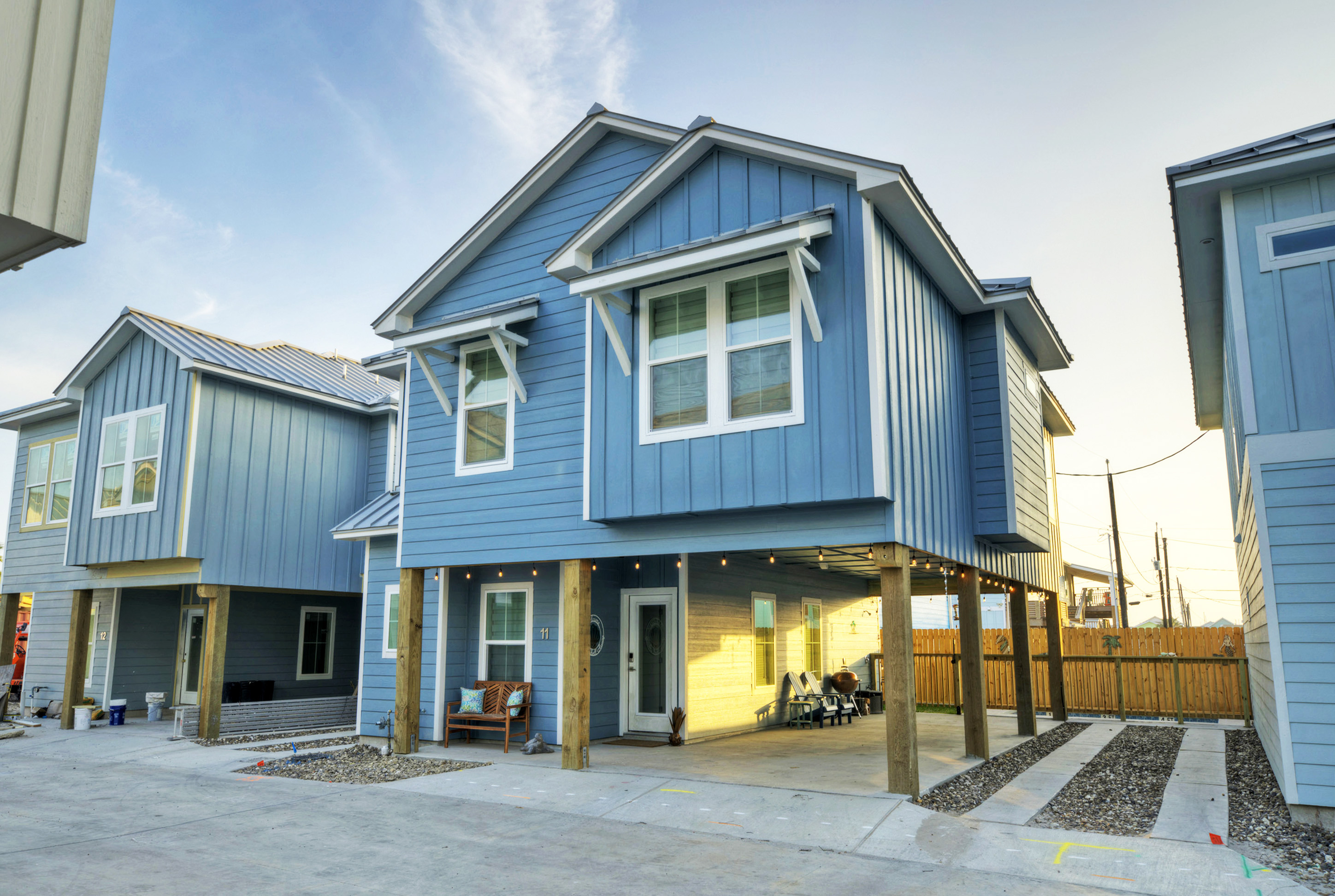 A modern blue two-story house with a metal roof, situated on stilts. The house features large windows and a covered porch area with seating. Gravel landscaping surrounds the base of the house.