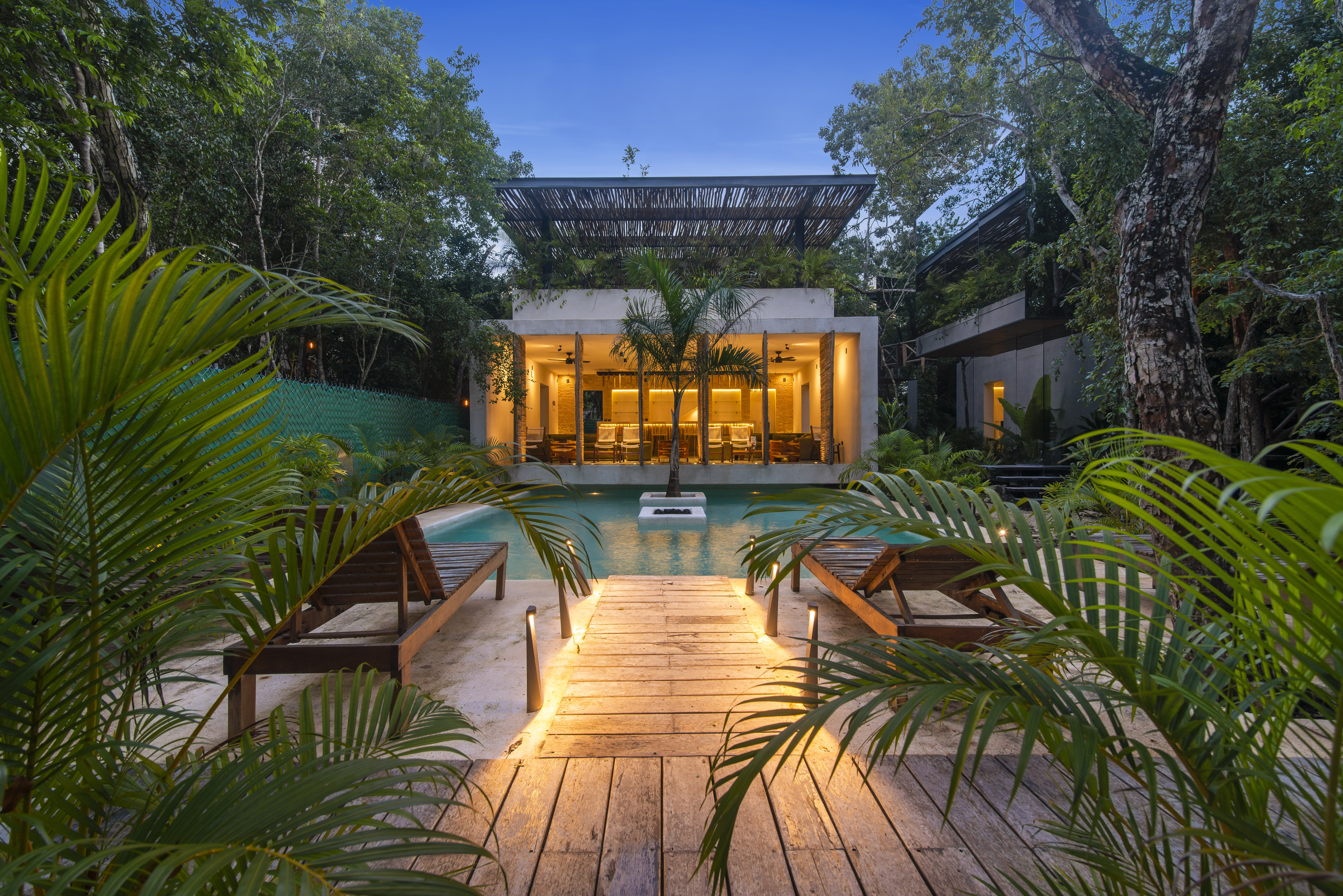 A serene outdoor space featuring a modern home surrounded by lush greenery. In the foreground, wooden lounge chairs are positioned beside a swimming pool, with a wooden walkway illuminated by soft lights leading towards the house. The house has large glass windows that showcase warm interior lighting, and a palm tree is centrally located in the pool area, enhancing the tranquil ambiance of the scene. The sky is a soft blue, suggesting twilight.
