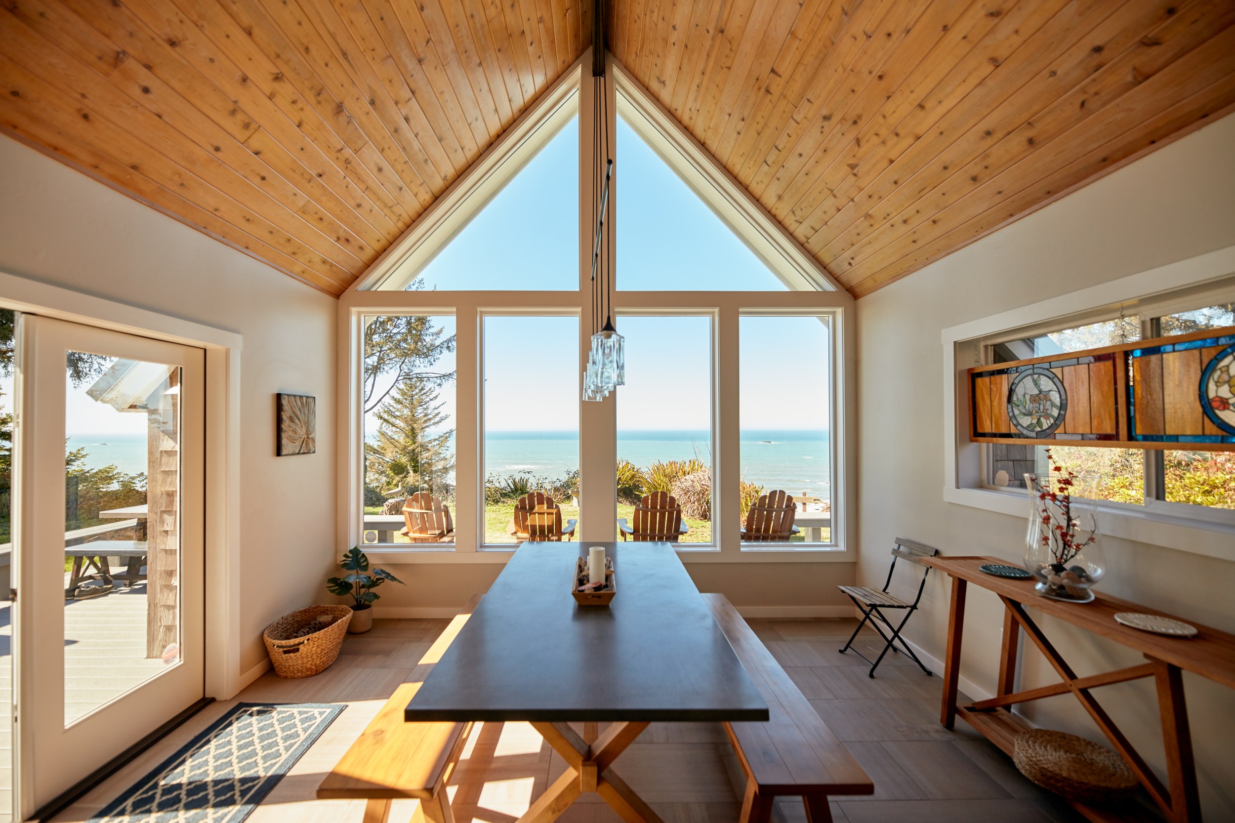 Dining room with a high wooden ceiling, large windows, and sea view. Features a long table, benches, decorative items, and stained glass accents.