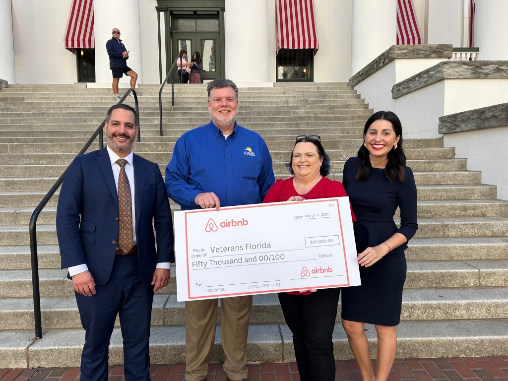 Group of four people standing at the bottom of a set of stairs holds a jumbo check from Airbnb addressed to Veterans Florida for $50,000.
