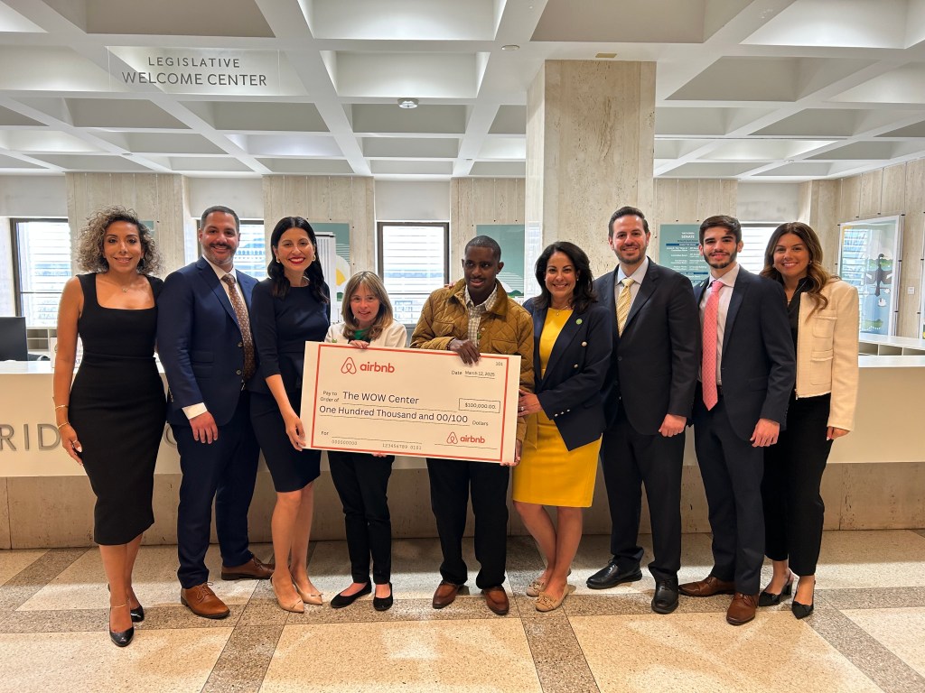 Group of nine people stand inside of a building holding a jumbo check from Airbnb addressed to The WOW Center for $100,000