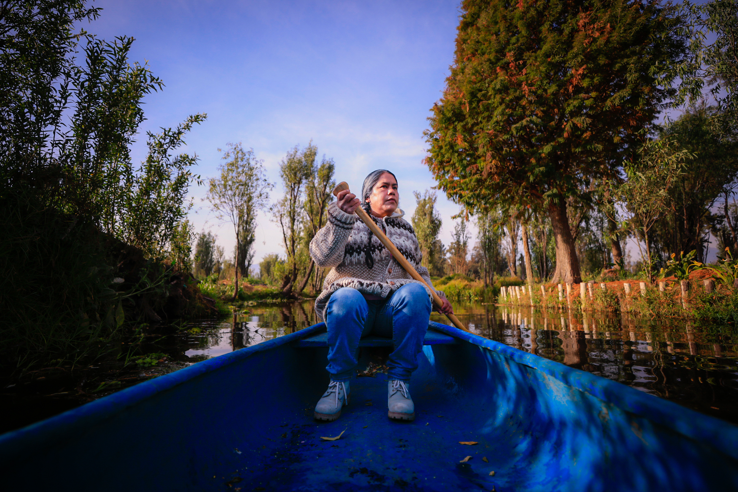 Rosalba Person paddling a blue canoe through a serene tree-lined canal at sunset.