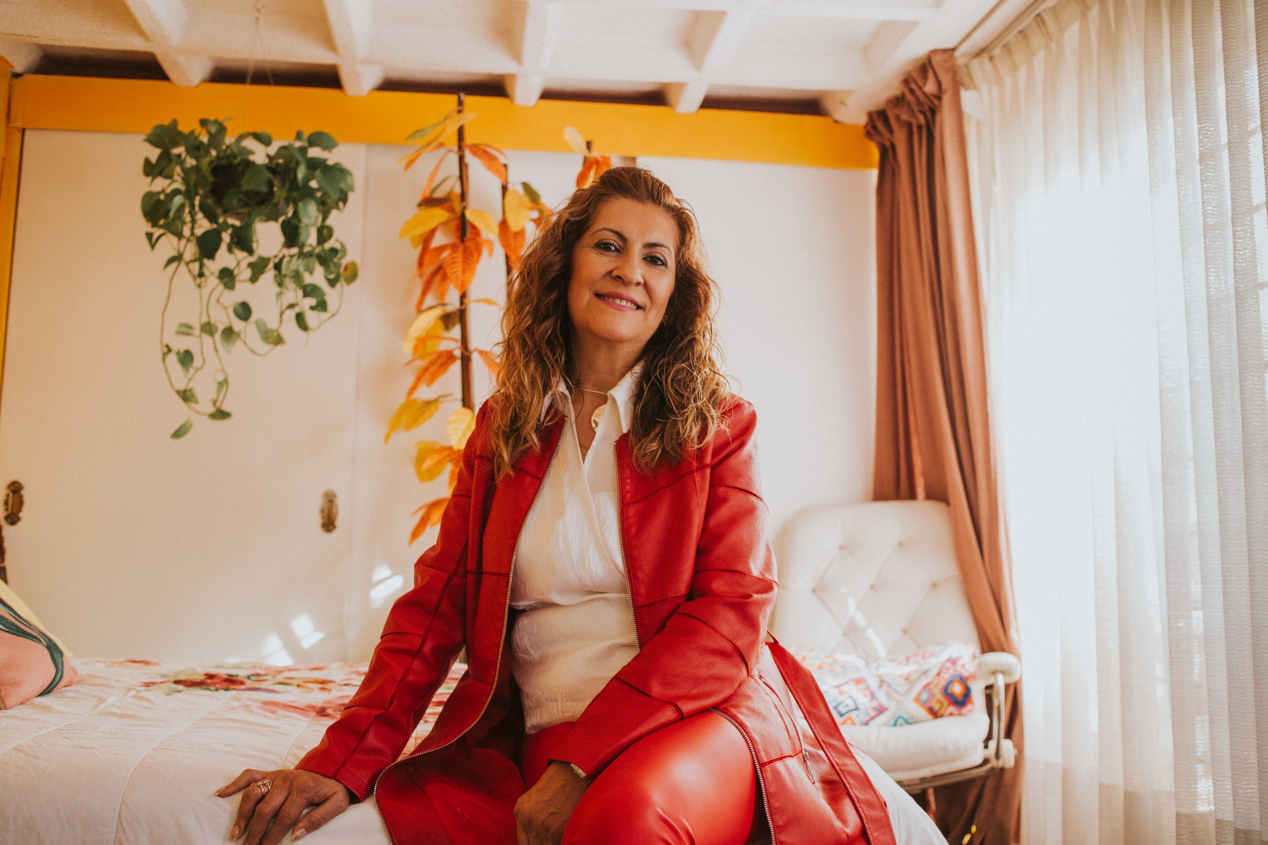 Diana Rios Person in a red outfit sitting on a bed in a sunlit room with plants and curtains.