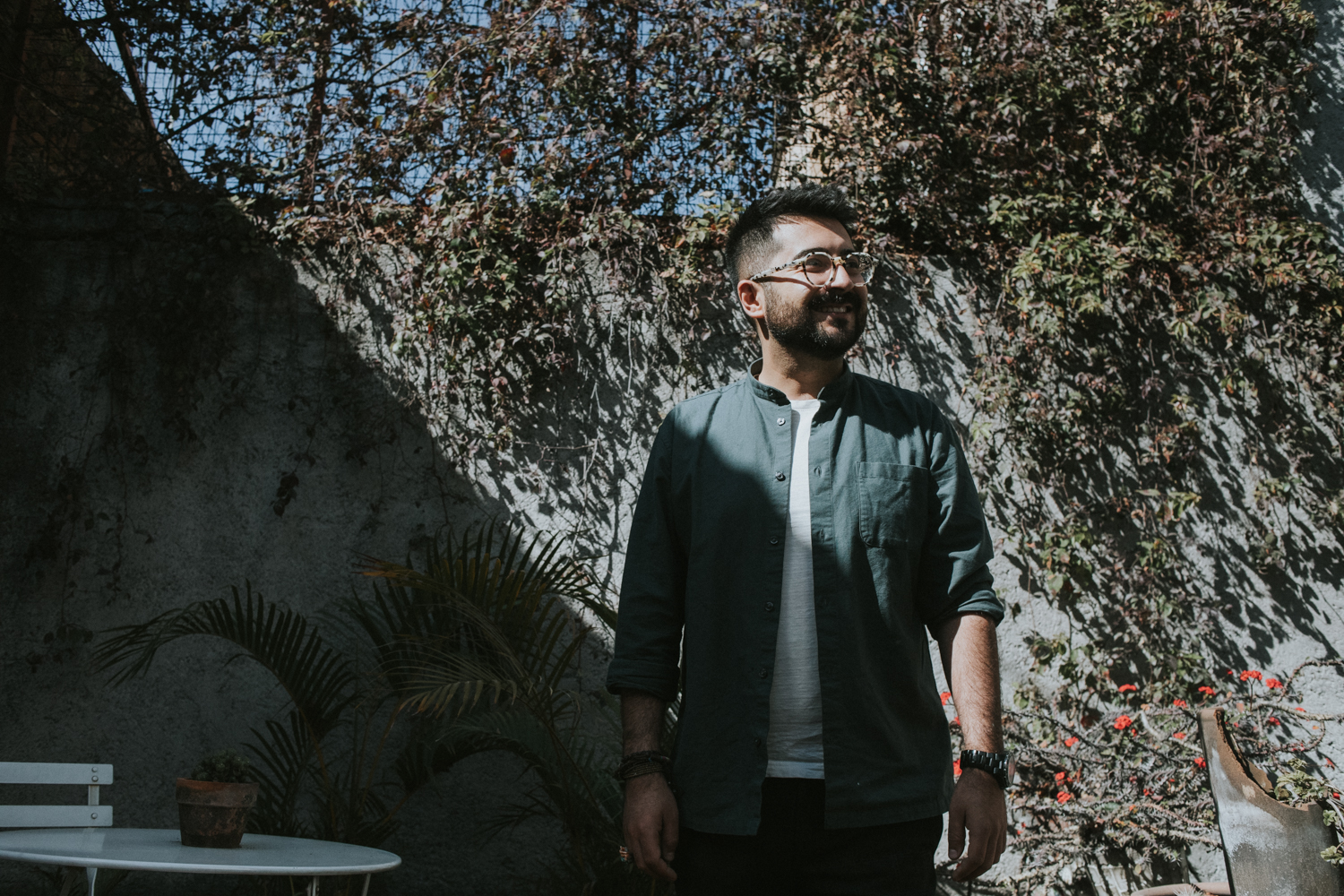 Person standing in sunlit garden with ivy-covered wall and white chair nearby