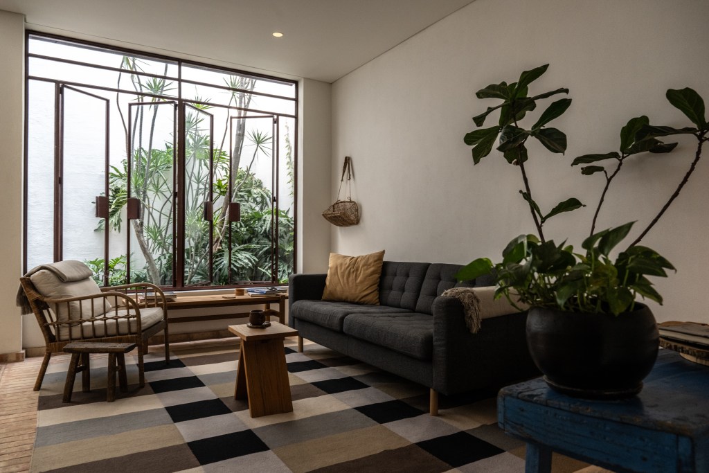 Cozy living room with a gray couch and wooden chairs, overlooking a lush garden through large windows.