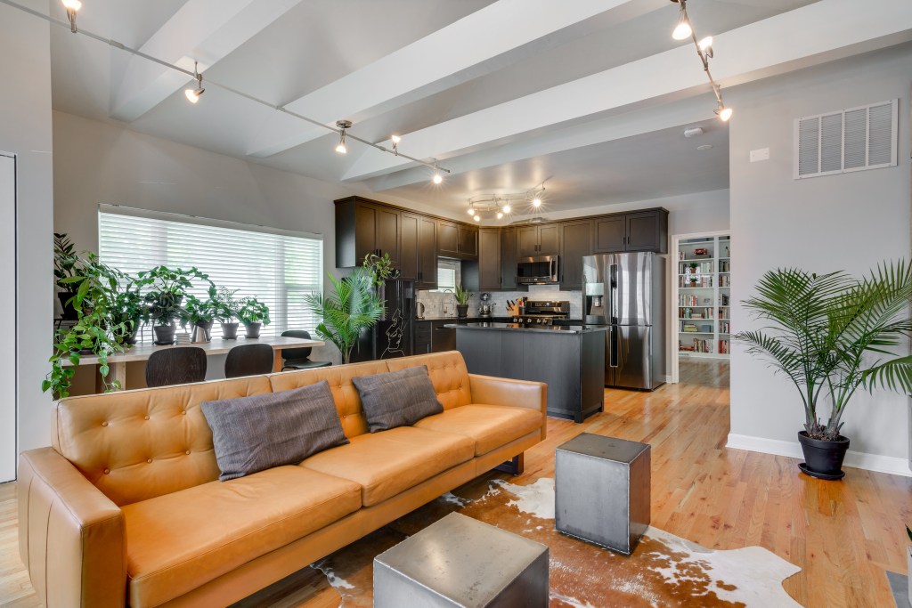 Spacious modern living room with an orange leather sofa, wooden floors, and a kitchen in the background featuring stainless steel appliances and dark cabinetry.