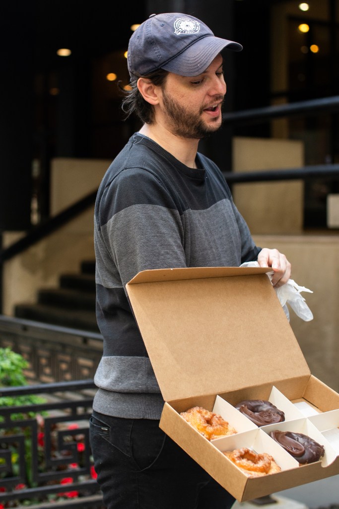 A person holding an open box of assorted donuts, including glazed and chocolate-covered varieties, standing outdoors near a building staircase.