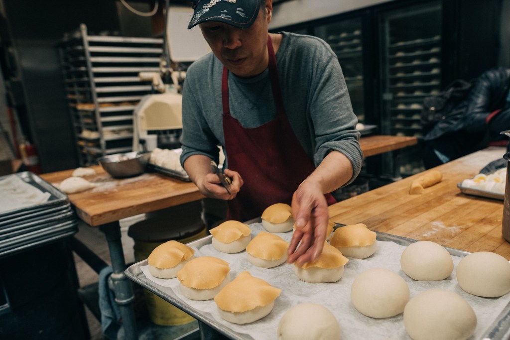 A baker in a red apron adds finishing touches to freshly baked bread rolls on a tray in a bakery kitchen, surrounded by wooden countertops and baking equipment.