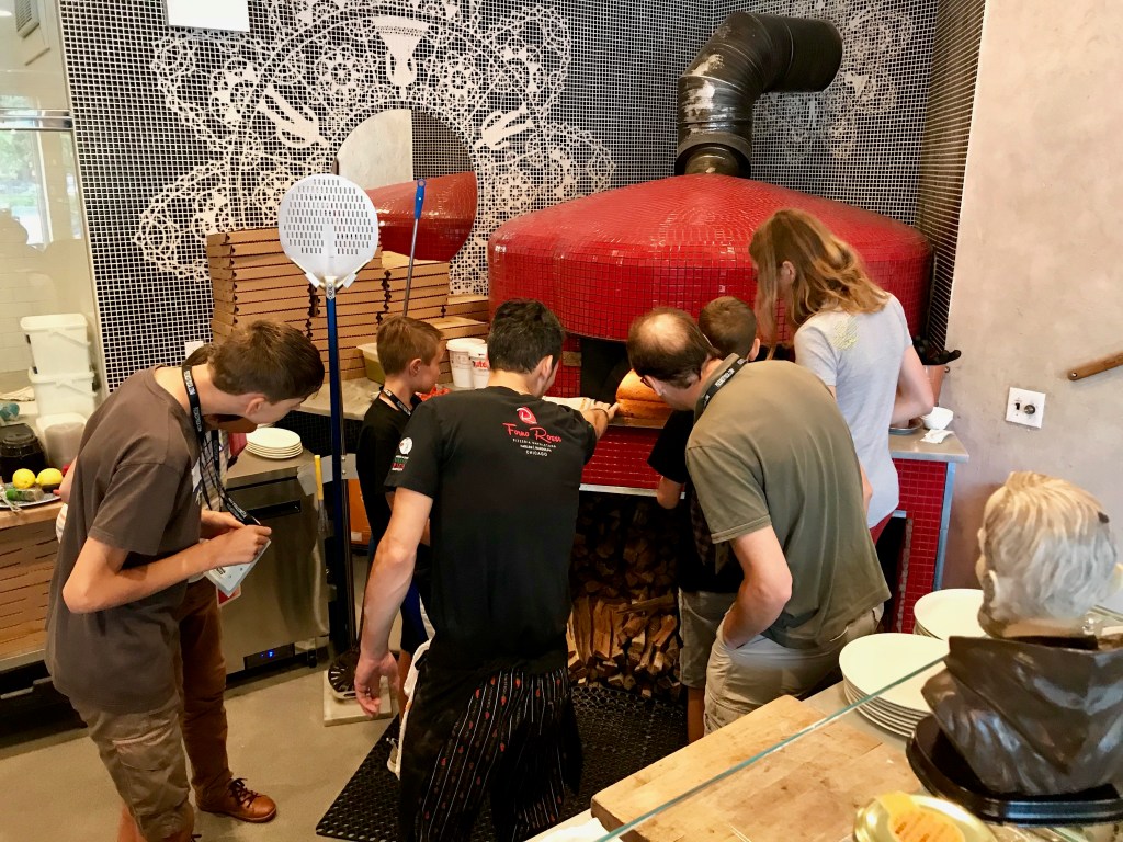 Group of people gathered around a modern red pizza oven, observing as a pizza is placed inside, with wood stacked below the oven and intricate mosaic tile design on the walls.