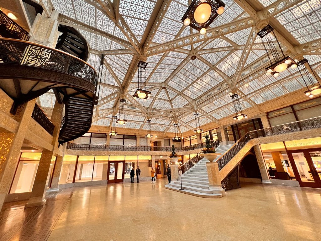 A spacious, sunlit atrium with intricate metalwork, a glass-paneled ceiling, and a grand staircase, showcasing elegant architectural design.