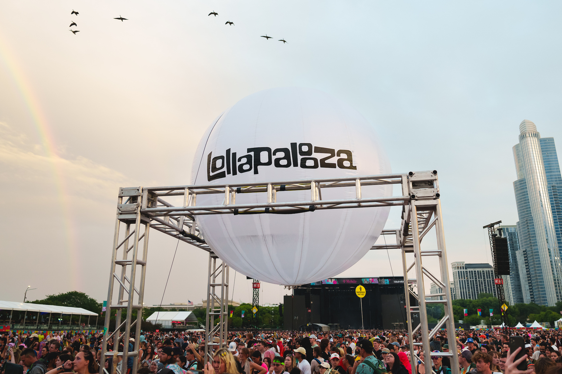 A large crowd gathered at the Lollapalooza festival beneath a white balloon with the Lollapalooza logo, with a city skyline and a rainbow visible in the background.