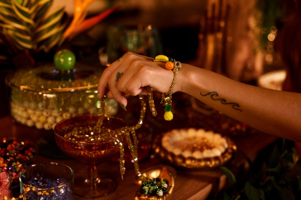 A close-up of a hand with wave tattoo and colorful bracelet holding gold beads over a festive table with bowls of decorative ornaments.