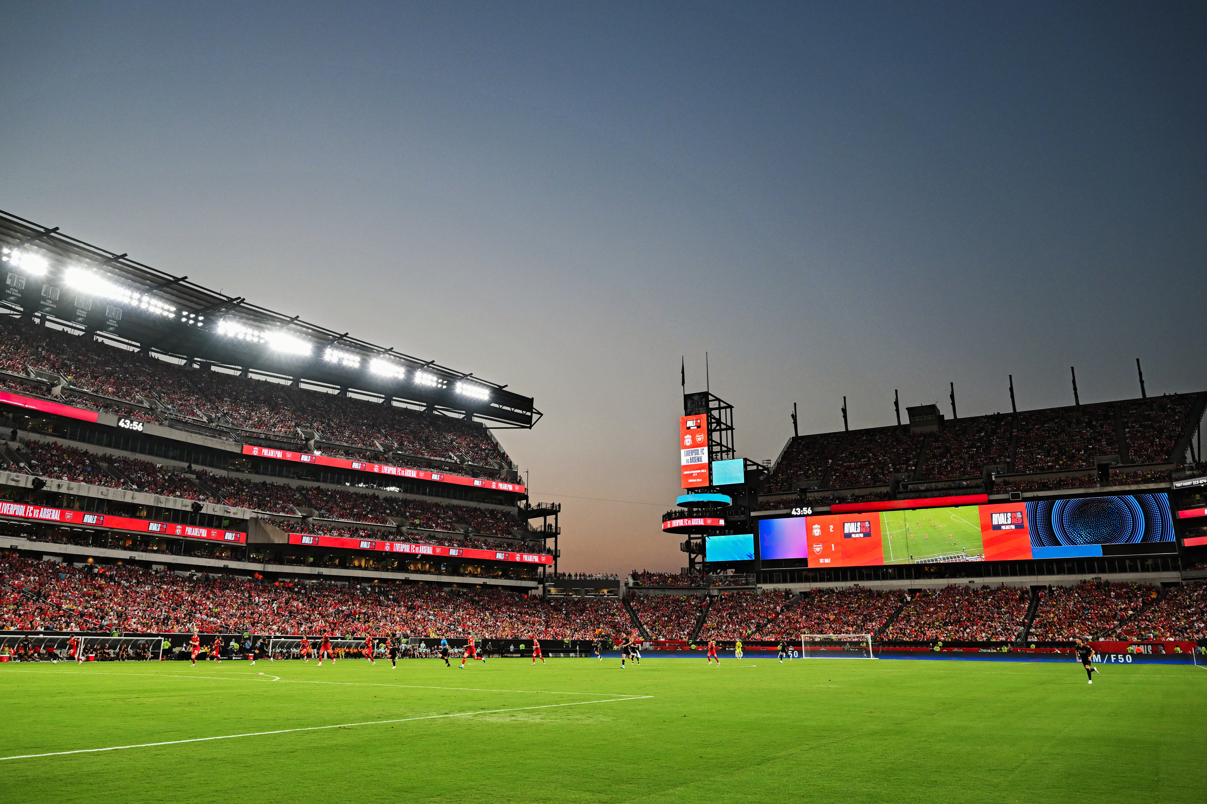 A stadium filled with spectators watching a soccer match at dusk, with brightly lit screens displaying scores.