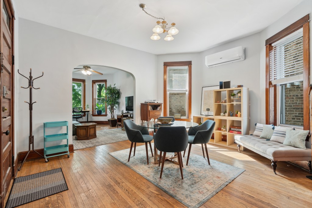 Bright, cozy dining area with four modern chairs around a wooden table, large windows letting sunlight in, and an adjoining living space with plants and a TV visible through an arched entryway.
