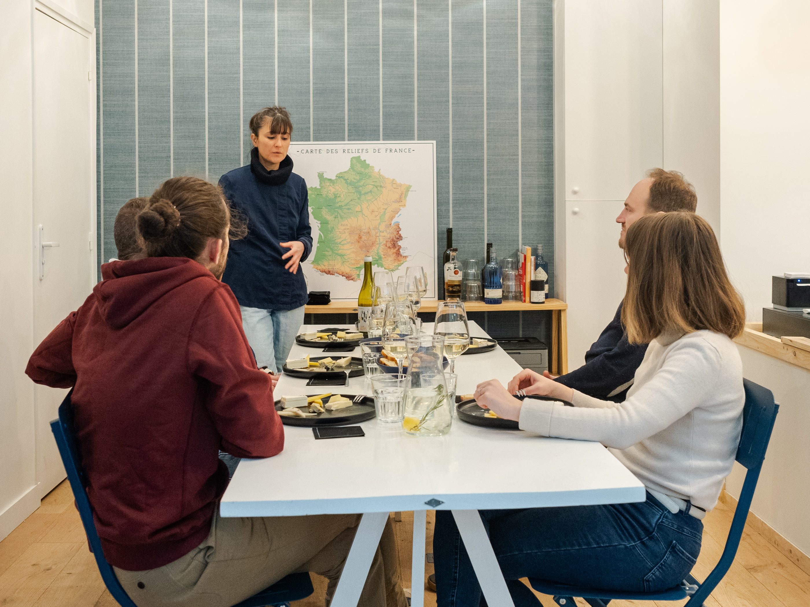 Several people attend an educational tasting or workshop at a table, listening to a presenter with a map of France in the background.