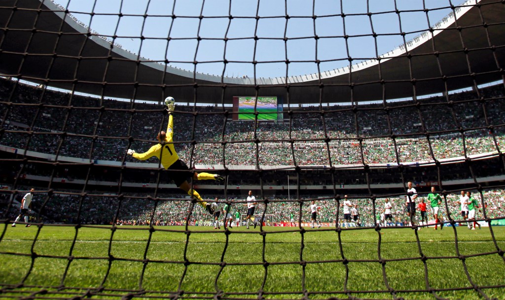 Goalkeeper in a yellow jersey leaping to save a soccer ball at a packed stadium.
