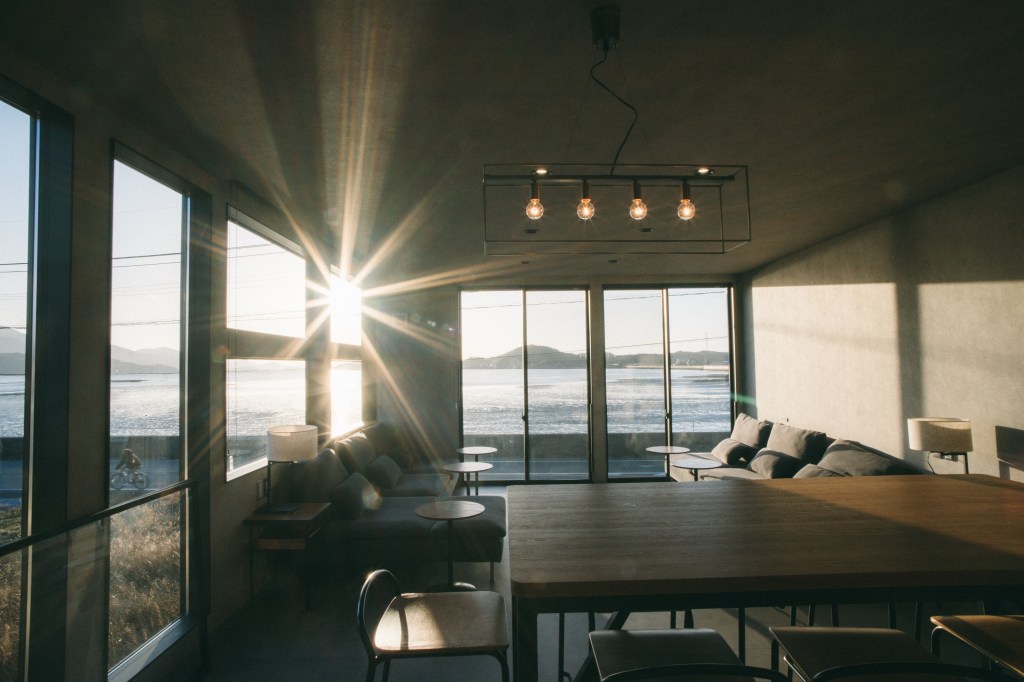 A modern, sunlit living space with large windows overlooking a serene waterfront and distant mountains during sunset.