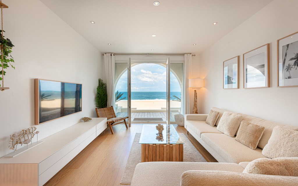 Bright and modern living room with beige furniture, large glass doors opening to a beachfront view with blue skies and ocean waves.