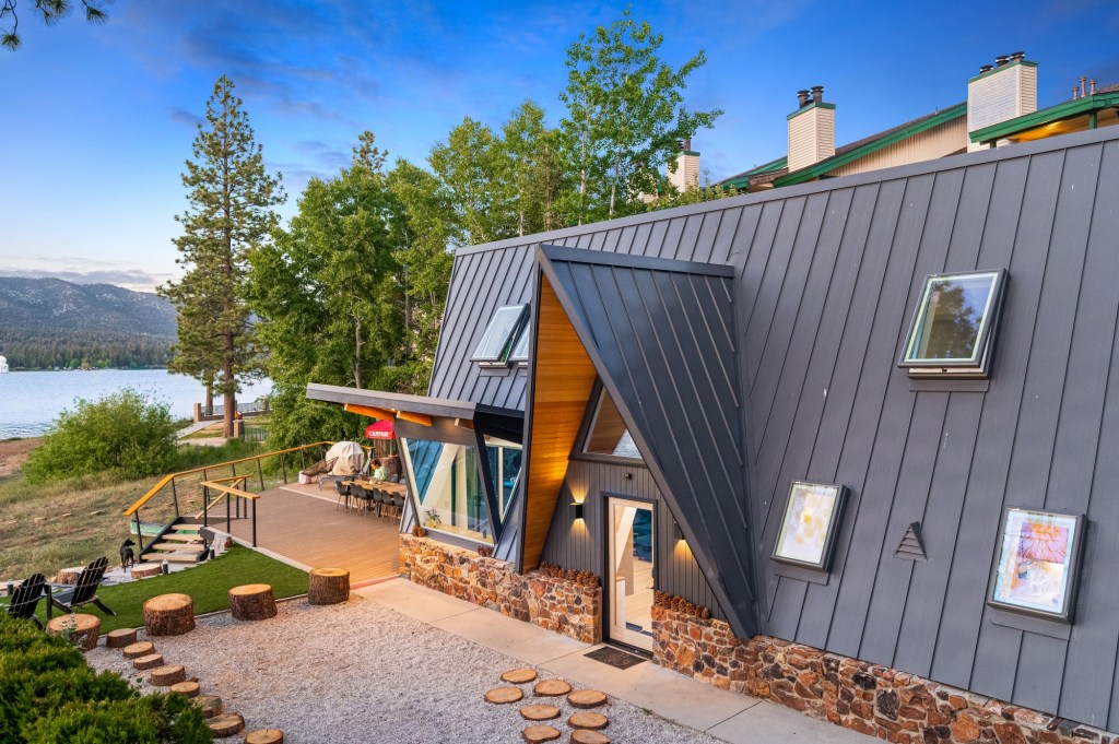 A modern A-frame cabin with gray metal siding, large triangular windows, and a wooden deck overlooking a serene lake surrounded by trees during sunset.