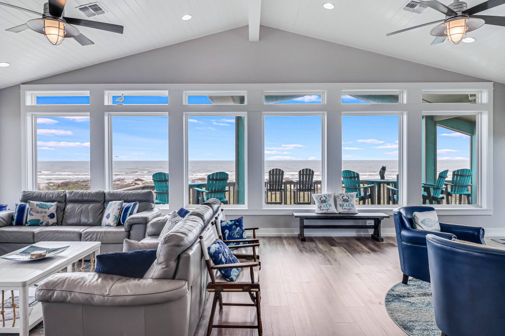 Spacious living room with coastal-themed decor, large windows displaying ocean views, and outdoor seating on a balcony with blue Adirondack chairs.