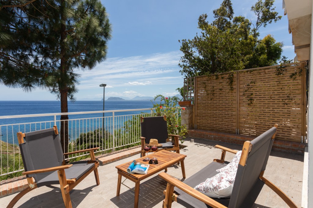 Sunny balcony with lounge chairs and a table set for relaxation, offering a picturesque view of the sea, framed by pine trees and a bamboo privacy fence.