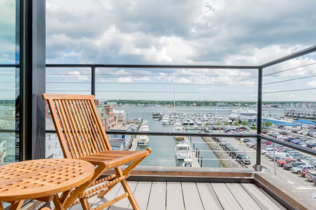 A private balcony with a wooden chair and table overlooking a marina filled with boats under a partly cloudy sky.