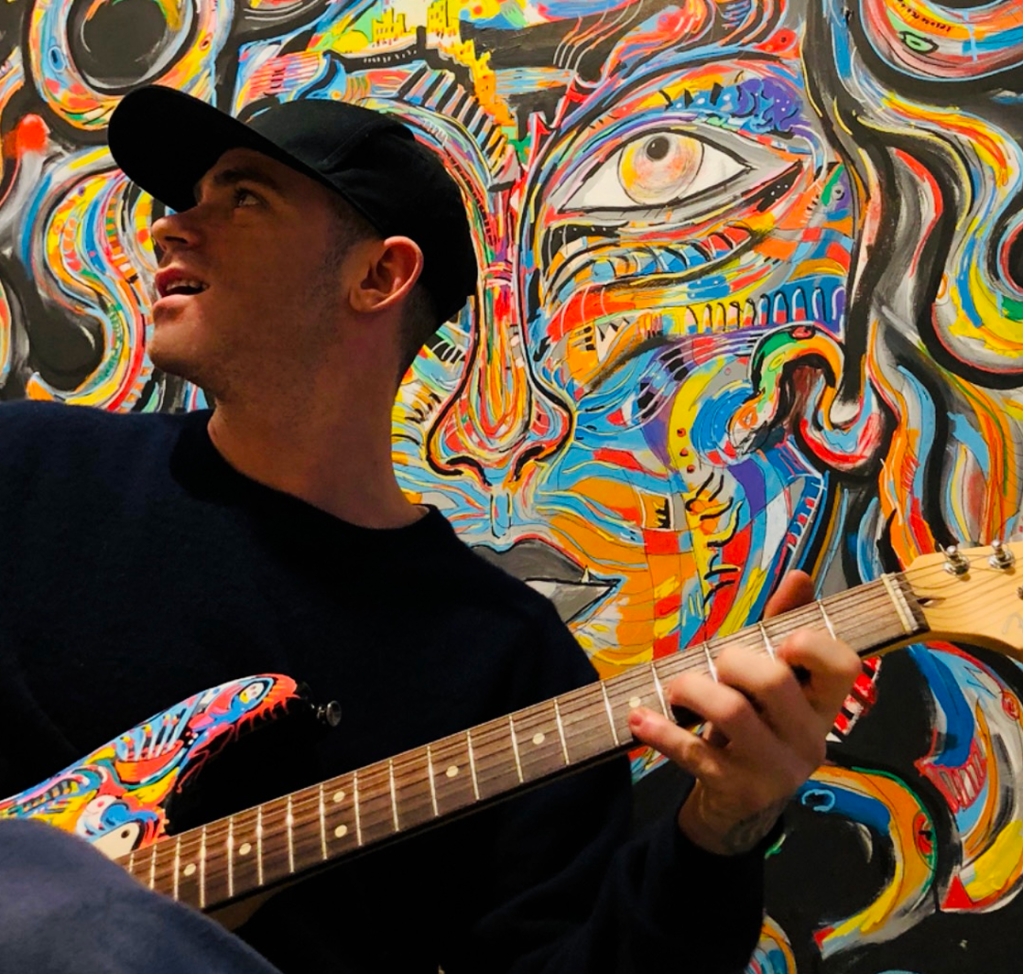 A person plays a vibrantly painted guitar in front of a colorful, abstract mural.