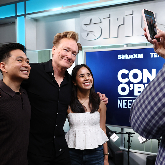 A SiriusXM studio setting with people posing for a picture, featuring a sign that reads 'CONAN O'BRIEN...' in the background.