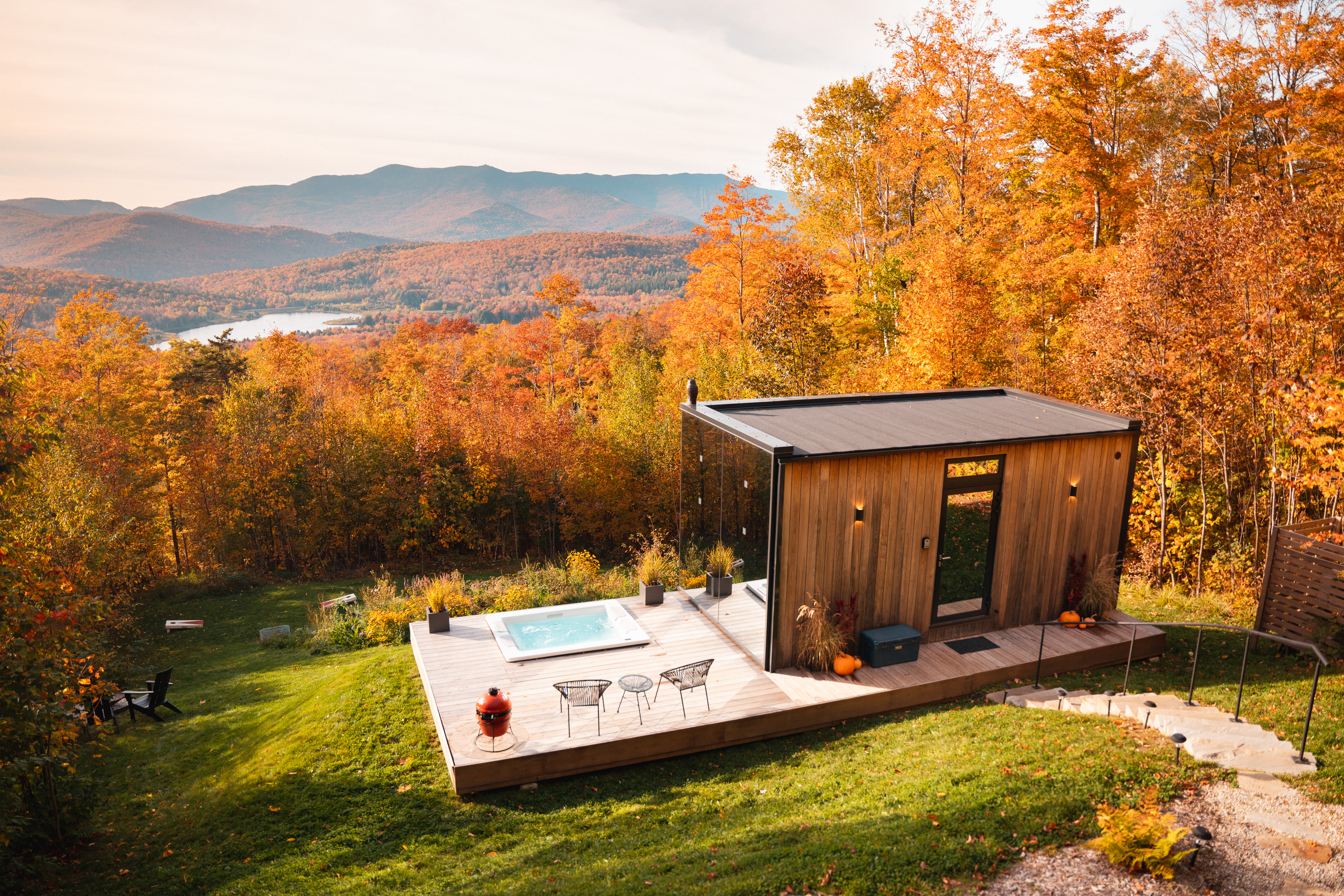 A modern wooden cabin with a deck and hot tub sits amidst vibrant autumn foliage overlooking a scenic mountain and lake view.