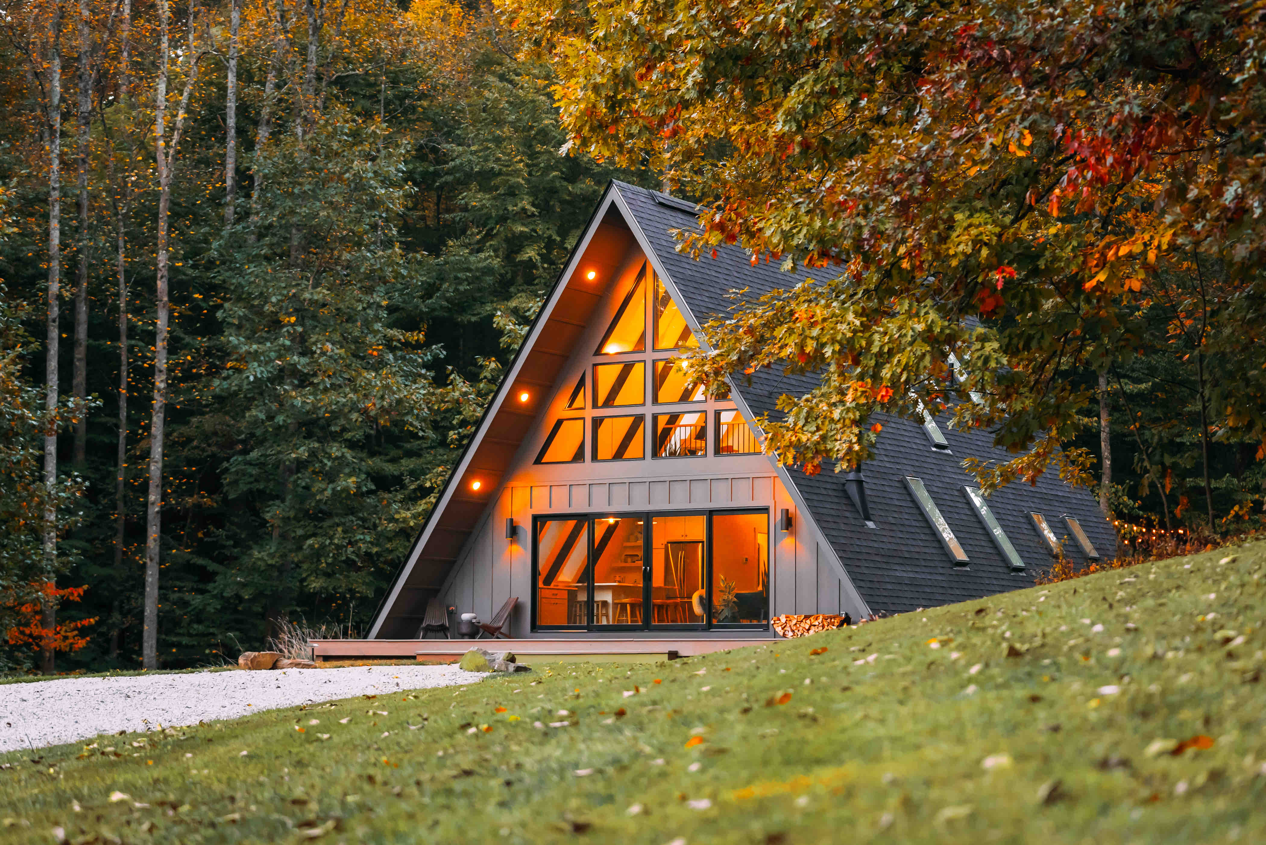 A warmly lit A-frame house with large windows stands on a gentle grassy slope, framed by colorful autumn trees.