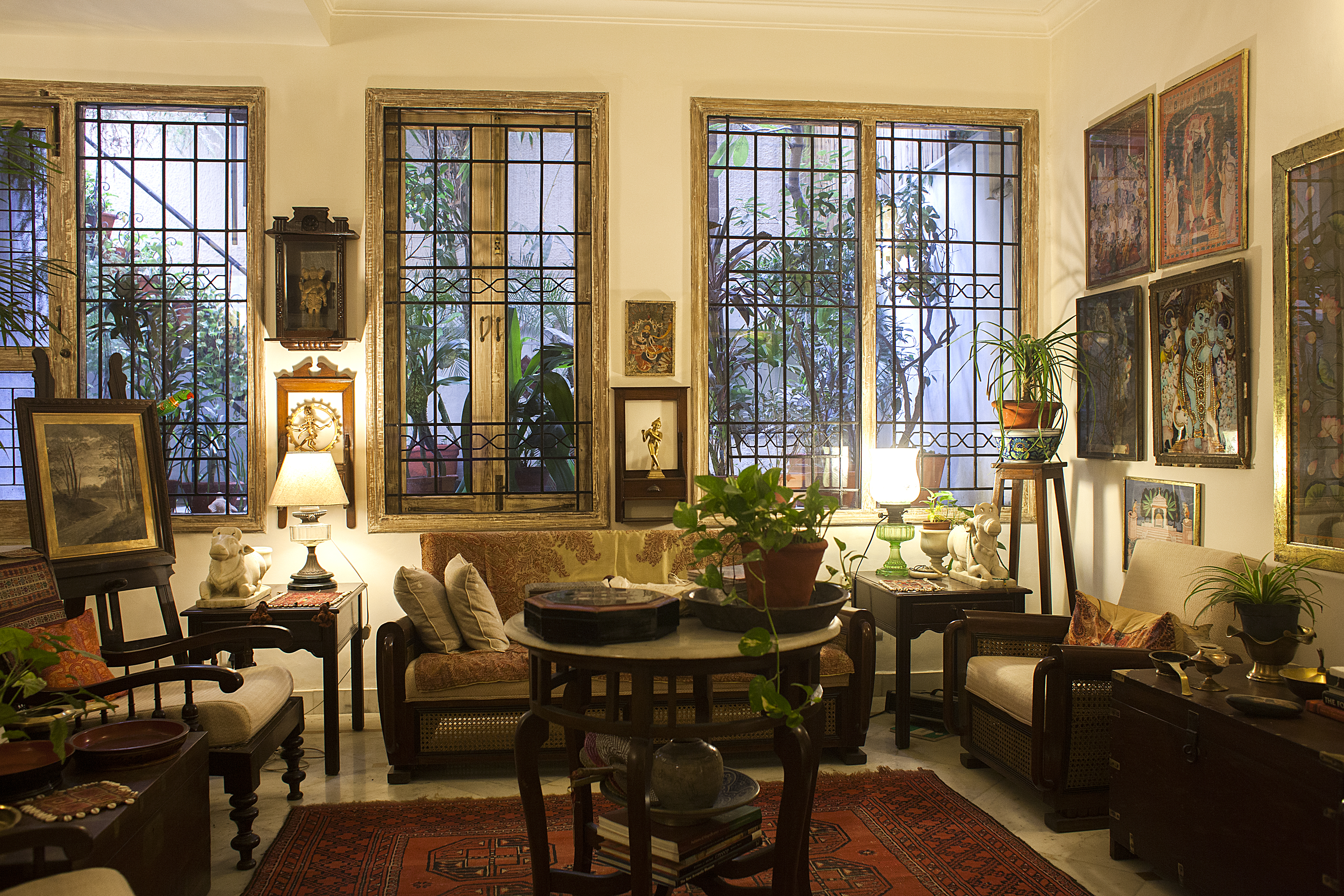 Cozy living room with warm lighting, featuring vintage furniture, framed artwork on walls, and potted plants near large windows with decorative iron grills.