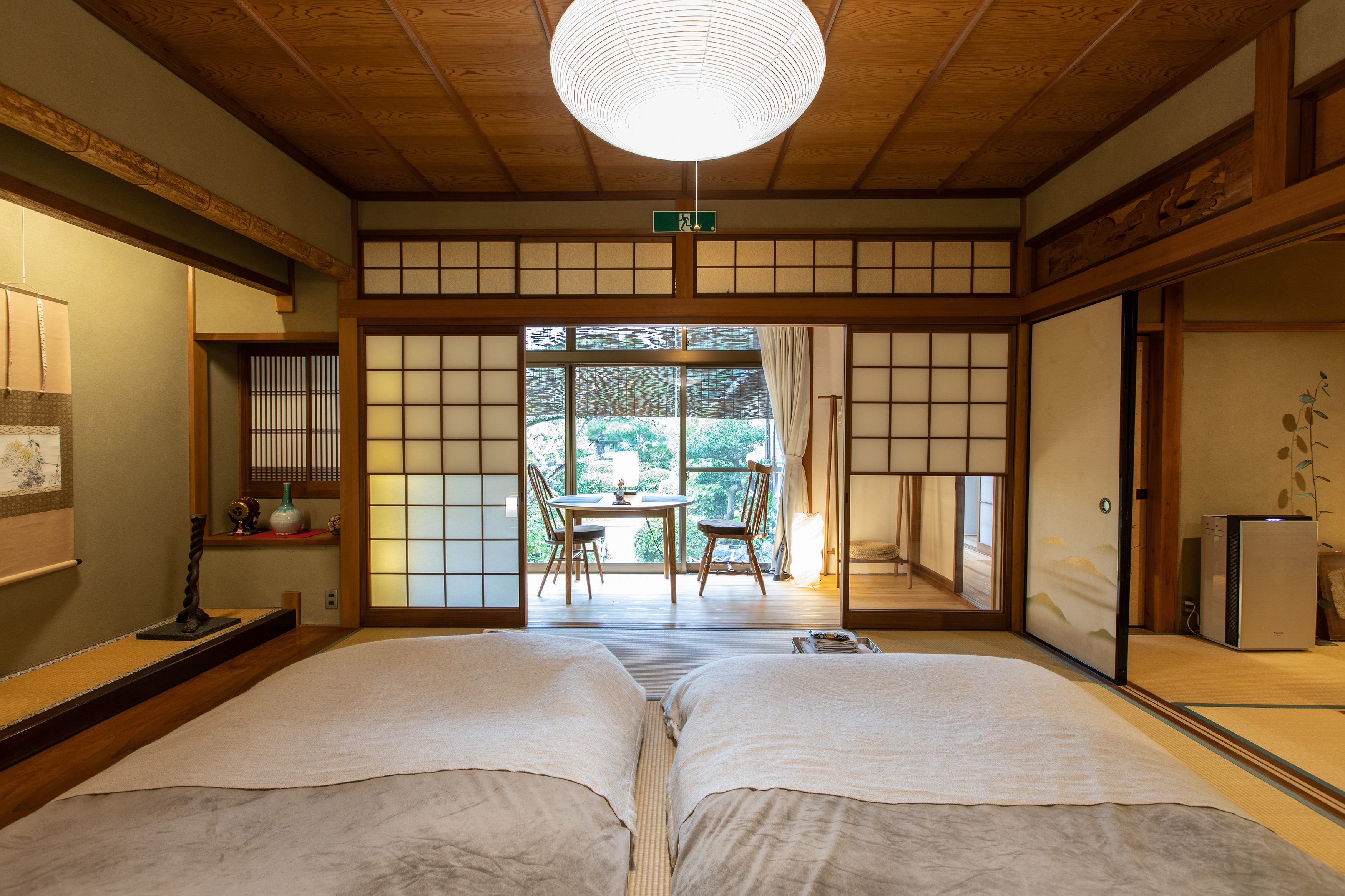 Traditional Japanese tatami room featuring futon bedding, shoji sliding doors, and a bright, peaceful garden view with a table and chairs in a sunlit area beyond.