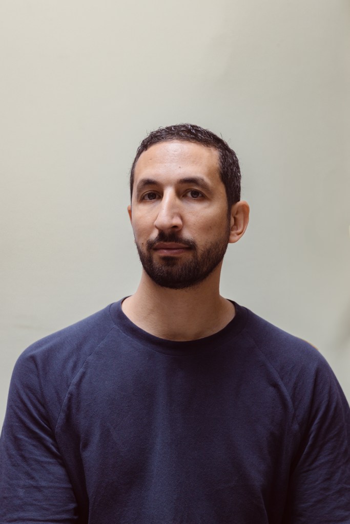 Individual in a plain navy blue shirt, standing against a simple beige backdrop.