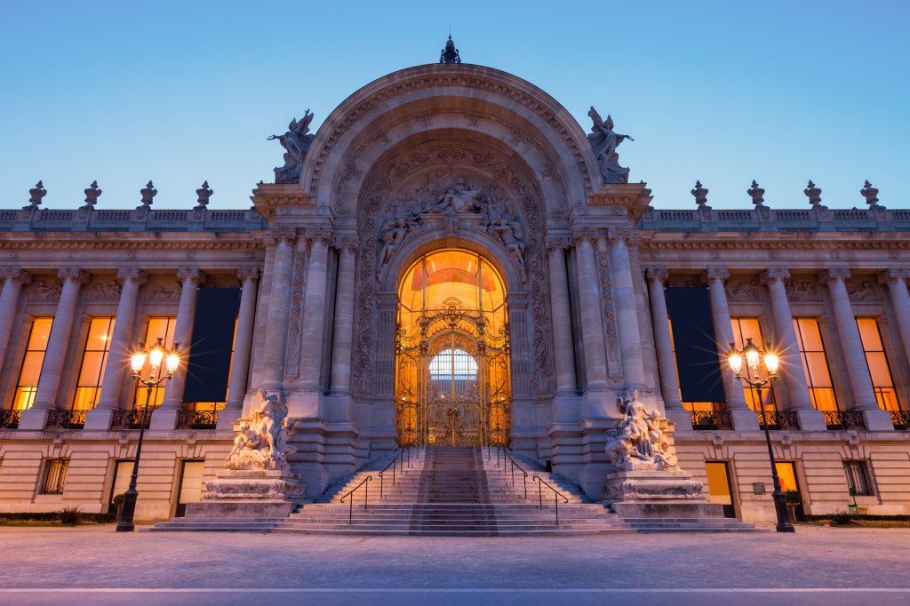 The grand entrance of the Petit Palais in Paris, illuminated at dusk with warm orange lights and intricate architectural details.