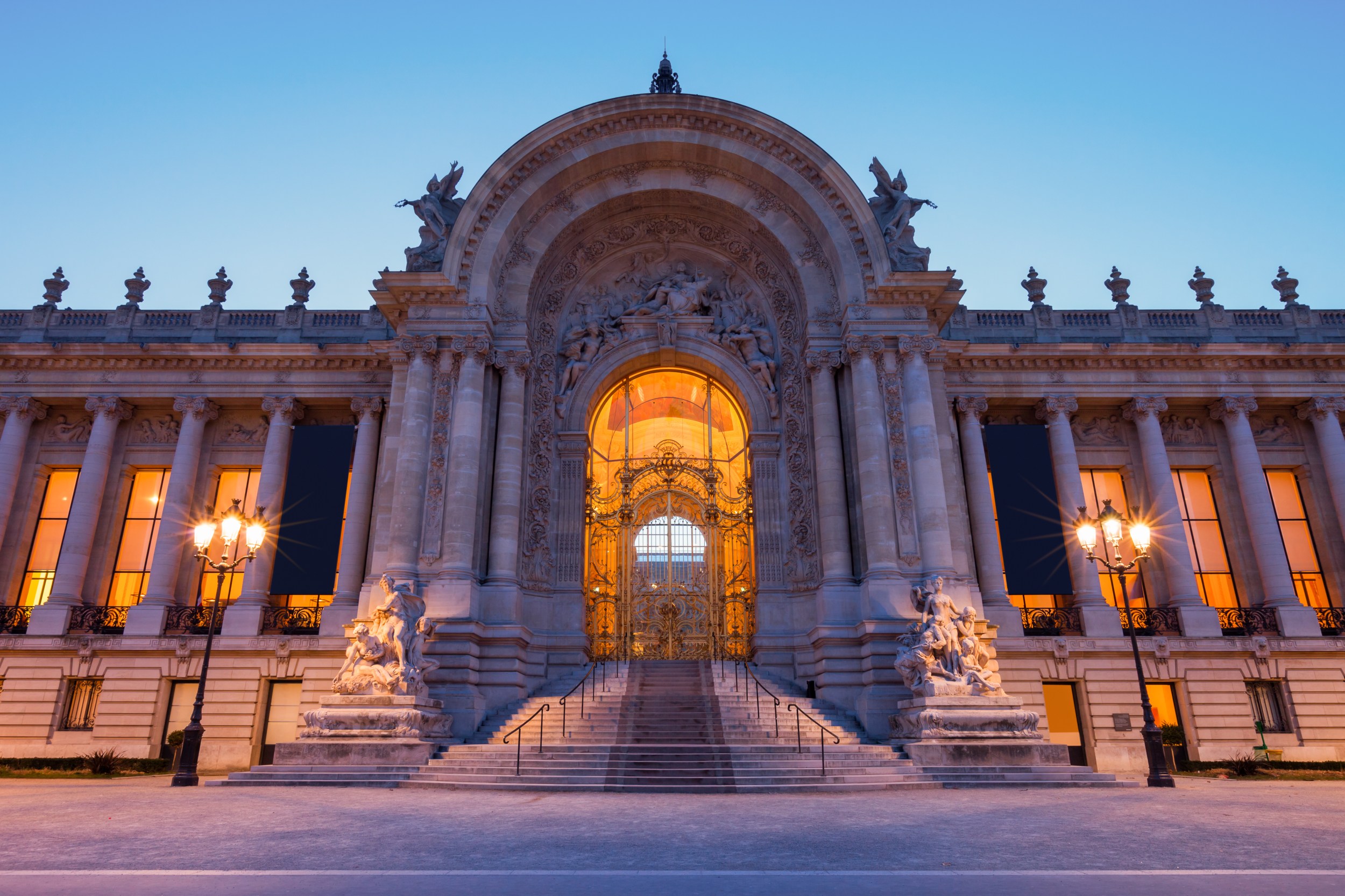 The grand entrance of the Petit Palais in Paris, illuminated at dusk with warm orange lights and intricate architectural details.