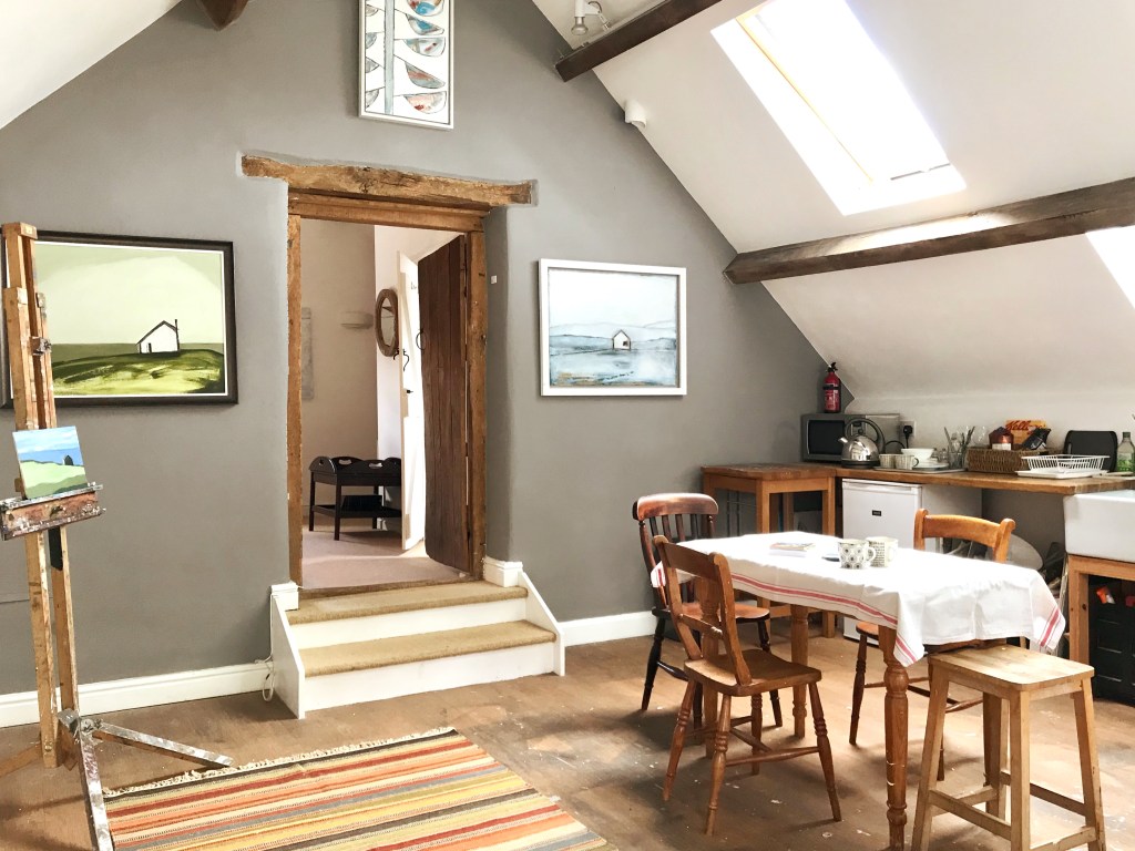 Bright artist's studio with skylights, a dining table, and paintings on the walls, featuring a cozy and rustic design with wooden beams and an easel displaying a landscape painting.
