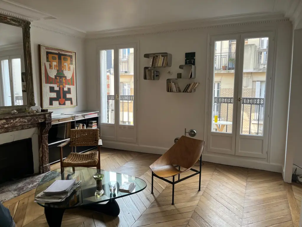 An elegant Parisian room boasts large windows, chevron wood floors, a marble fireplace, and minimalist modern furniture for a sophisticated look.