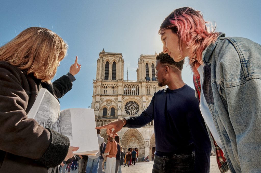A group gathers in front of Notre Dame, discussing its architecture and history with the help of a guidebook and enthusiastic gestures.
