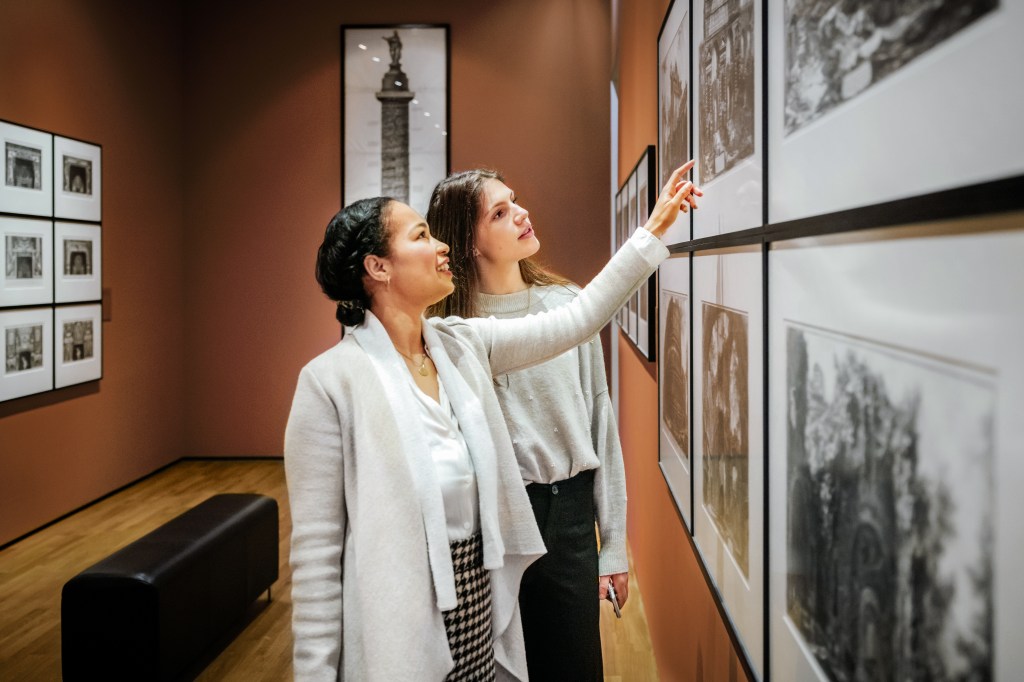 Two people in a warmly lit gallery space examining framed artwork on a wall, with a focus on detailed black-and-white illustrations.