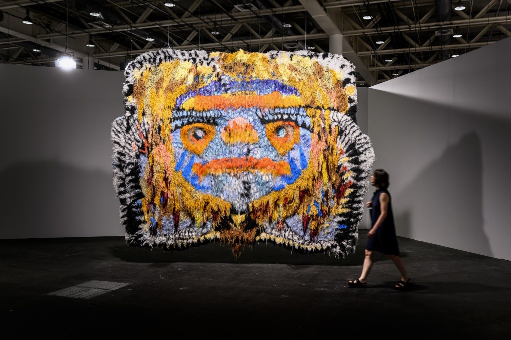 An abstract artwork with vivid colors and a symmetrical design resembling a face, featuring orange, blue, and yellow tones, showcased in a modern exhibit hall under bright lighting, with a visitor walking nearby.