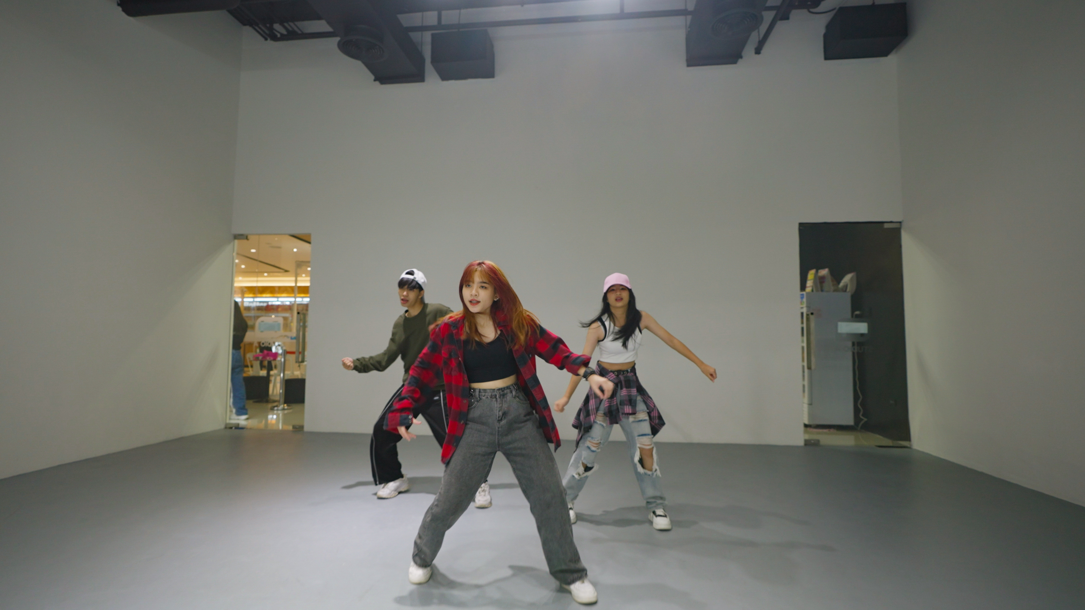 Three young women dancing in a brightly lit studio, wearing casual outfits with plaid shirts and sneakers.