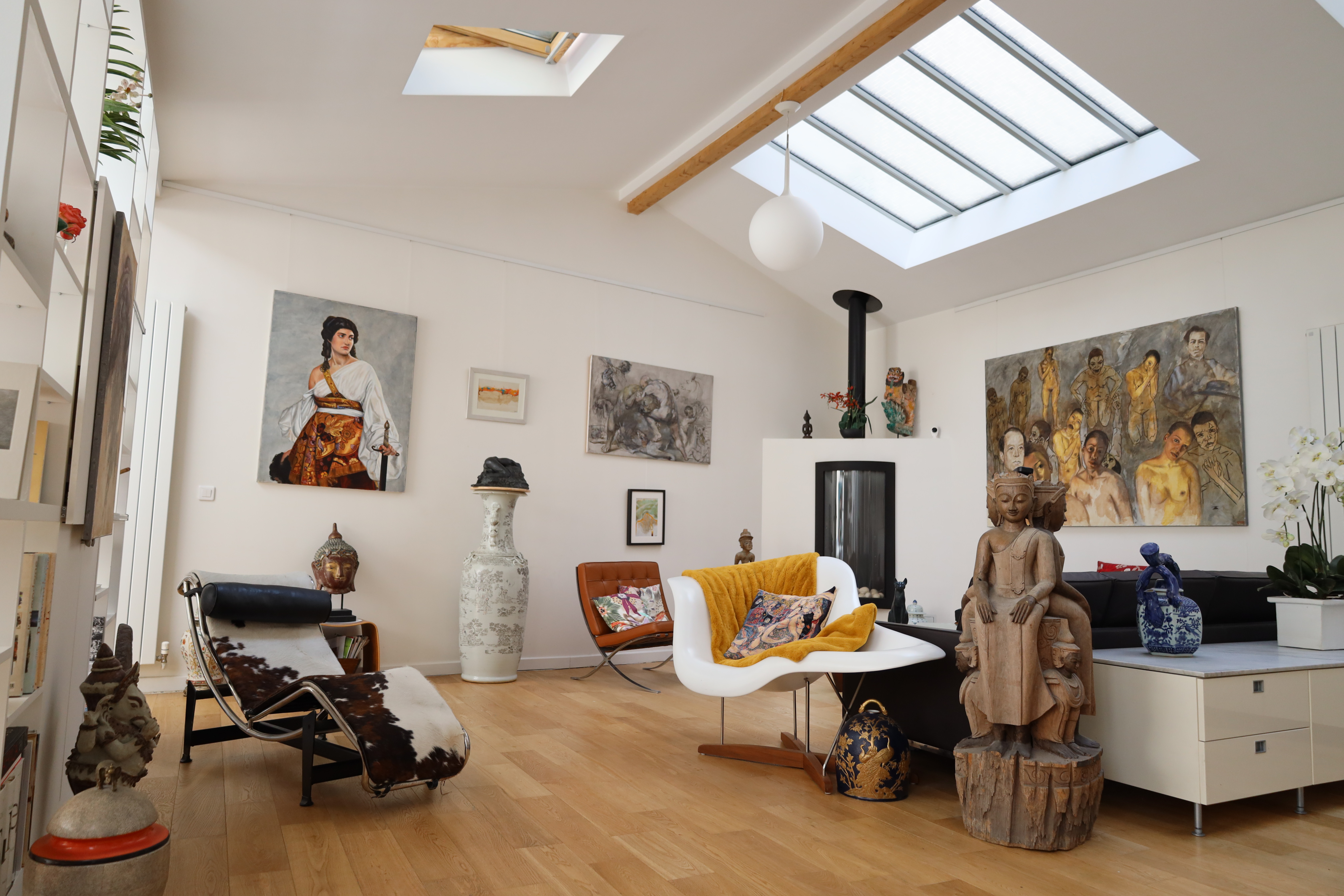 Spacious living room with modern and eclectic decor, featuring colorful artwork on the walls, a skylight, and wood flooring. A mix of furniture includes a sculpted wooden chair, contemporary seating, and decorative vases.