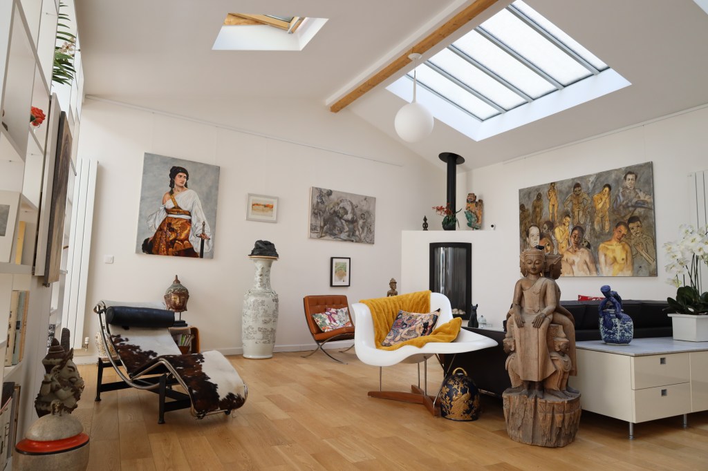 Spacious living room with modern and eclectic decor, featuring colorful artwork on the walls, a skylight, and wood flooring. A mix of furniture includes a sculpted wooden chair, contemporary seating, and decorative vases.